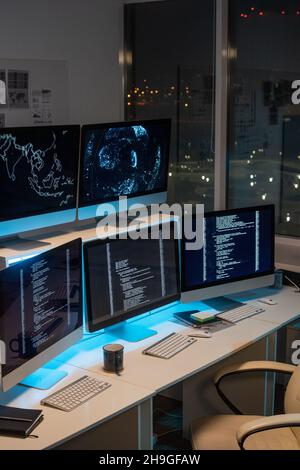 Group of computer monitors with coded data standing in row on desks in empty office of programming company at night time Stock Photo