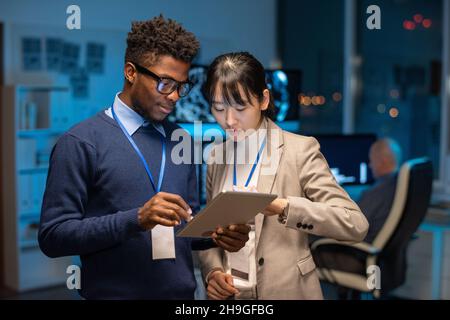 Two young interracial colleagues preparing presentation while scrolling through online information in tablet Stock Photo