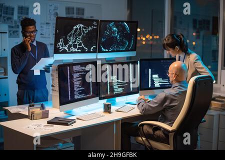 Two colleagues sitting in front of computer monitors with decoded data while their African coworker with paper speaking on mobile phone Stock Photo