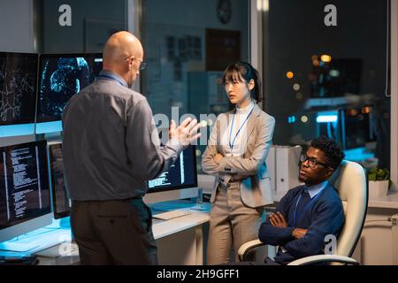 Mature manager giving instructions to young intercultural subordinates at working meeting late at night Stock Photo