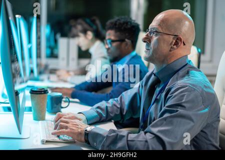 Serious mature businessman looking at computer screen while typing against two intercultural coworkers Stock Photo