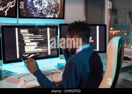 Young tired African businessman sitting in front of computer monitor with coded data on screen while working overtime Stock Photo
