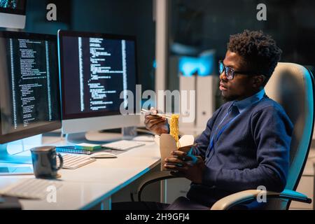 Young hungry programmer eating chinese wok in front of computer screen with coded data while working in office at night Stock Photo