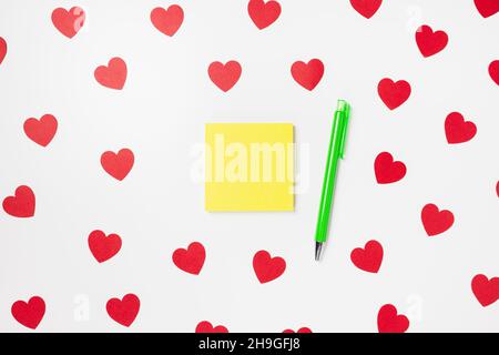 Self-stick yellow note pad or sticky note and pen on white with red hearts Valentines Day background. Shopping gifts, to do list concept, copy space, mockup for message Stock Photo