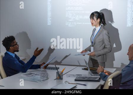 Young African businessman talking to Asian female colleague standing by large interactive whiteboard Stock Photo