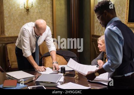 Three intercultural lawyers working with papers at meeting while two men standing by table and looking through documents Stock Photo