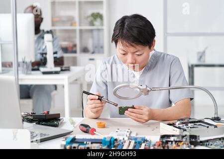 Asian woman in uniform soldering circuit board of electronic device while looking in magnifying glass Stock Photo