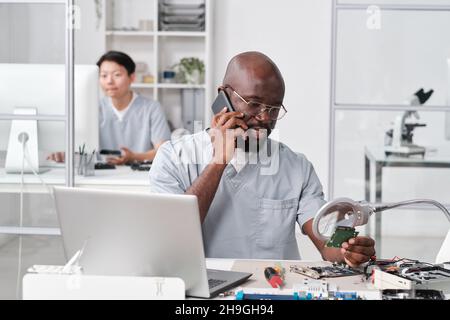 Young African repairman speaking on mobile phone to client in front of laptop in workshop Stock Photo
