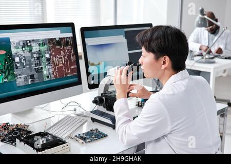Young contemporary researcher with microscope looking at computer screen during work in scientific laboratory Stock Photo