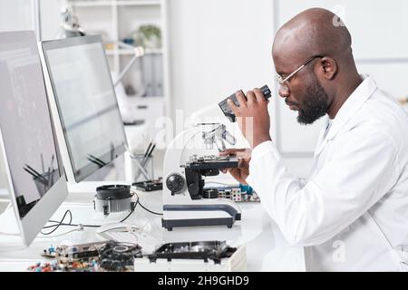 Side view of young African scientist in whitecoat studying microchip characteristics in microscope Stock Photo
