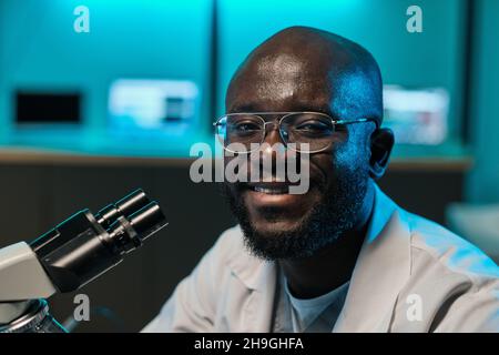Happy young African researcher in whitecoat looking at camera while sitting by microscope in scientific laboratory Stock Photo