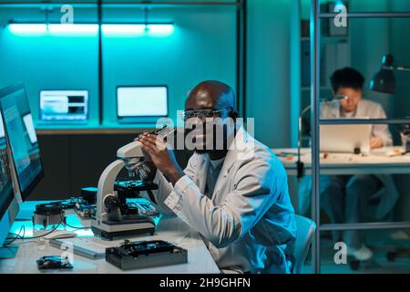 Young successful researcher with microscope sitting by workplace in scientific laboratory Stock Photo