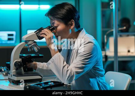 Young serious female laboratory worker in whitecoat looking at microchip in microscope while sitting by desk Stock Photo