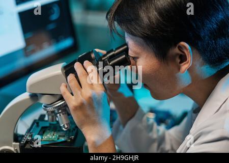 Young Asian technician looking in microscope while studying microchip characteristics in laboratory Stock Photo