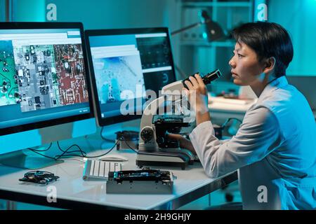 Young serious female scientist with microscope looking at computer screen while sitting by workplace Stock Photo