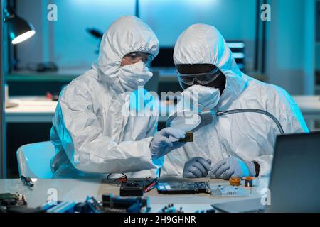 Young Asian woman in coveralls helping colleague with repairment of microchip of electronic device Stock Photo