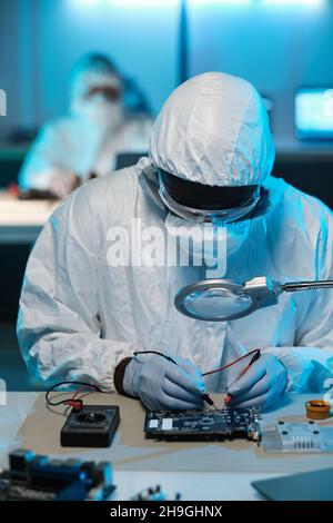 Contemporary repairman in protective workwear soldering circuit board while looking in magnifying glass Stock Photo