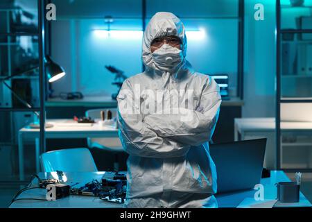 Young female in protective workwear standing against desk with laptop and hardware inside laboratory Stock Photo