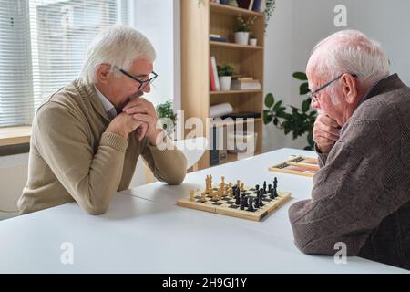 Elderly man in eyeglasses thinking about his chess move while sitting at the table and playing together with his friend Stock Photo
