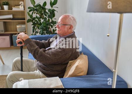 Side view of elderly man in eyeglasses resting on the sofa in the room leaning on the crutch Stock Photo