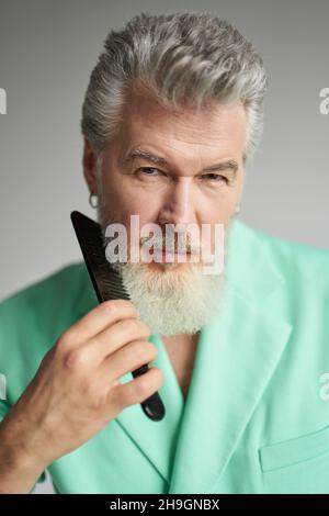Portrait of attractive gray haired mature man looking at camera, brushing his beard with comb, posing over white background Stock Photo