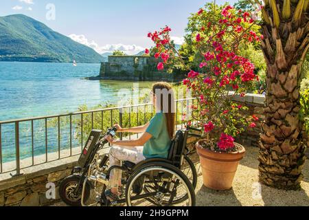 Young woman in wheelchair enjoying view on lakeside and mountains - Junge Rollstuhlfahrerin geniesst die Aussicht am Seeufer auf See und Berge Stock Photo