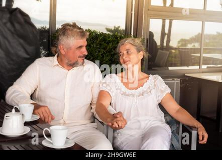 the couple of retirees sit together to the table on the veranda and drink tea, Stock Photo