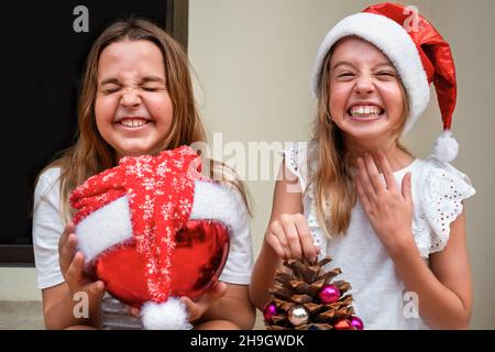 children girls in santa hats sit on the porch with Christmas decorations. children holding pine cone and christmas ornament Stock Photo