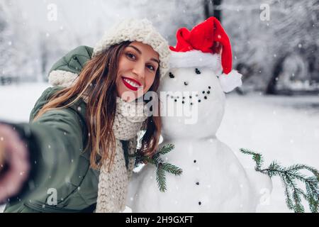 Happy woman takes selfie by snowman in Santa hat outdoors in snowy winter park. Christmas festive season. Fun activities Stock Photo