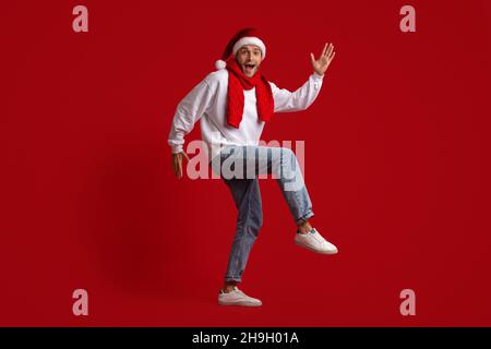 Christmas Dance. Funny Guy In Santa Hat Dancing Over Red Studio Background Stock Photo