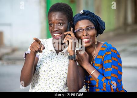 Two beautiful happy adult businesswomen talking on mobile phone and pointing finger in the street. Stock Photo