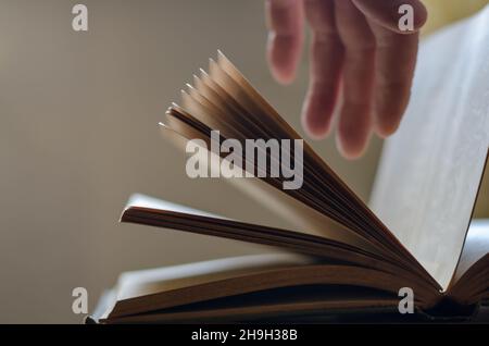 A defocused shot of an old open book and a man's hand. Palm hanging over top of dusty pages of hardcover book. Side view. Inside the room. Stock Photo