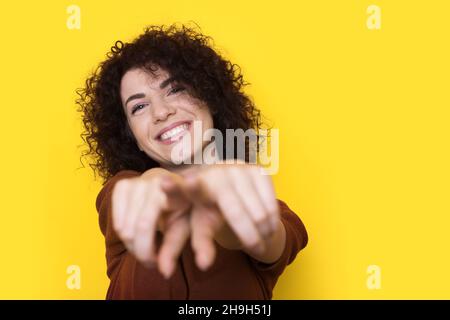 Portrait of a caucasian woman looking camera while pointing to you standing isolated over yellow background. Smiling curly haired woman. Stock Photo