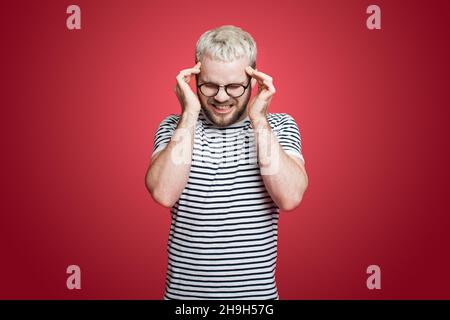 Portrait of a man pressing fingers to temples, closing eyes. Portrait of a man wearing eyeglasses isolated on red background suffering from severe Stock Photo