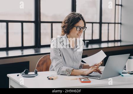 Focused European business woman sitting at home office holds financial documents, working on laptop Stock Photo