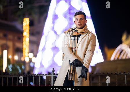 Young man adjusting his gloves at night with scarf in winter. Christmas tree in the background. Stock Photo