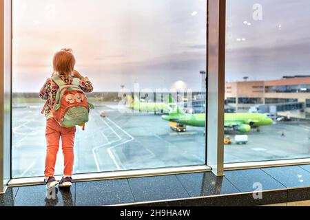 A little todd girl with a backpack looks out the panoramic windows at the planes. A tourist trip. An airport. Stock Photo