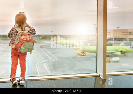 A little todd girl with a backpack looks out the panoramic windows at the planes. A tourist trip. An airport. Stock Photo