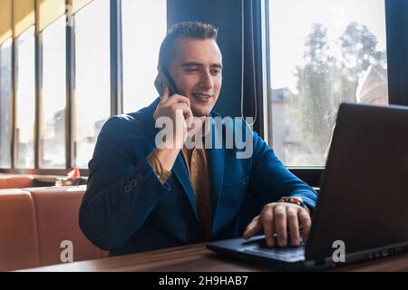 A business man stylish positive smiling businessman in an attractive European-looking suit works in a laptop, talks on a cell or mobile phone, sitting Stock Photo