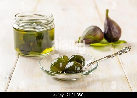 Delicious conserved unripe figs on a glass saucer, whole green figs in syrup jam jar and ripe fruits on a white wooden table. Traditional tasty jam. Stock Photo