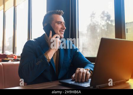 A business man stylish positive smiling businessman in an attractive European-looking suit works in a laptop, talks on a cell or mobile phone, sitting Stock Photo