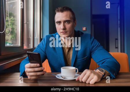 A seriously collected, brooding guy businessman of European appearance portrait sits at a table in a cafe on a coffee break and uses a smartphone or m Stock Photo