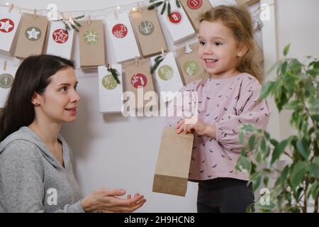Mother with child opening Christmas advent calendar tasks and gifts. Toddler girl excited about festive surprise in craft bag at home. Family Stock Photo