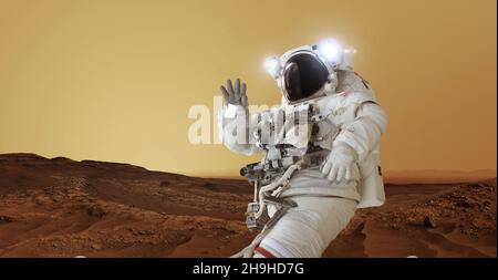 Astronaut in a space suit and a helmet with light stands on the red planet Mars greets and waves his hand. Welcome to Mars concept. Space man travels Stock Photo