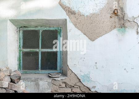 Small framed dusty window in a wall with disintegrated plastering showing layers of сement and brick. Dirty unwashed rectangular window. Stock Photo