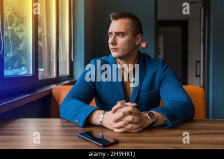 A brooding, stylish businessman portrait of European appearance in a jacket and shirt sits at a table in a cafe for a cup of coffee on a lunch break a Stock Photo