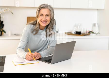 Cheerful mature female working remotely, business lady listening to an online webinar on laptop, senior woman taking notes in notebook, working on freelance project at home, e-learning concept Stock Photo