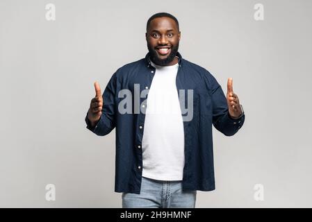 Positive African man with beard looks at camera, holding two hands in front of him showing the size on gray isolated background in studio, wearing casual shirt, smiling joyfully, presenting something Stock Photo