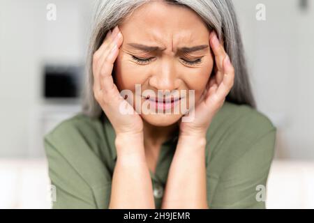 Middle-aged woman with long silver hair rubbing temples, suffering from migraine, mid-aged female feel strong painful headache, sitting with eyes closed and holding head Stock Photo