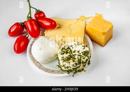 Types of cheese (sliced, hard, white with herbs) and heap of cherry tomato on plate on white table, closeup. Stock Photo